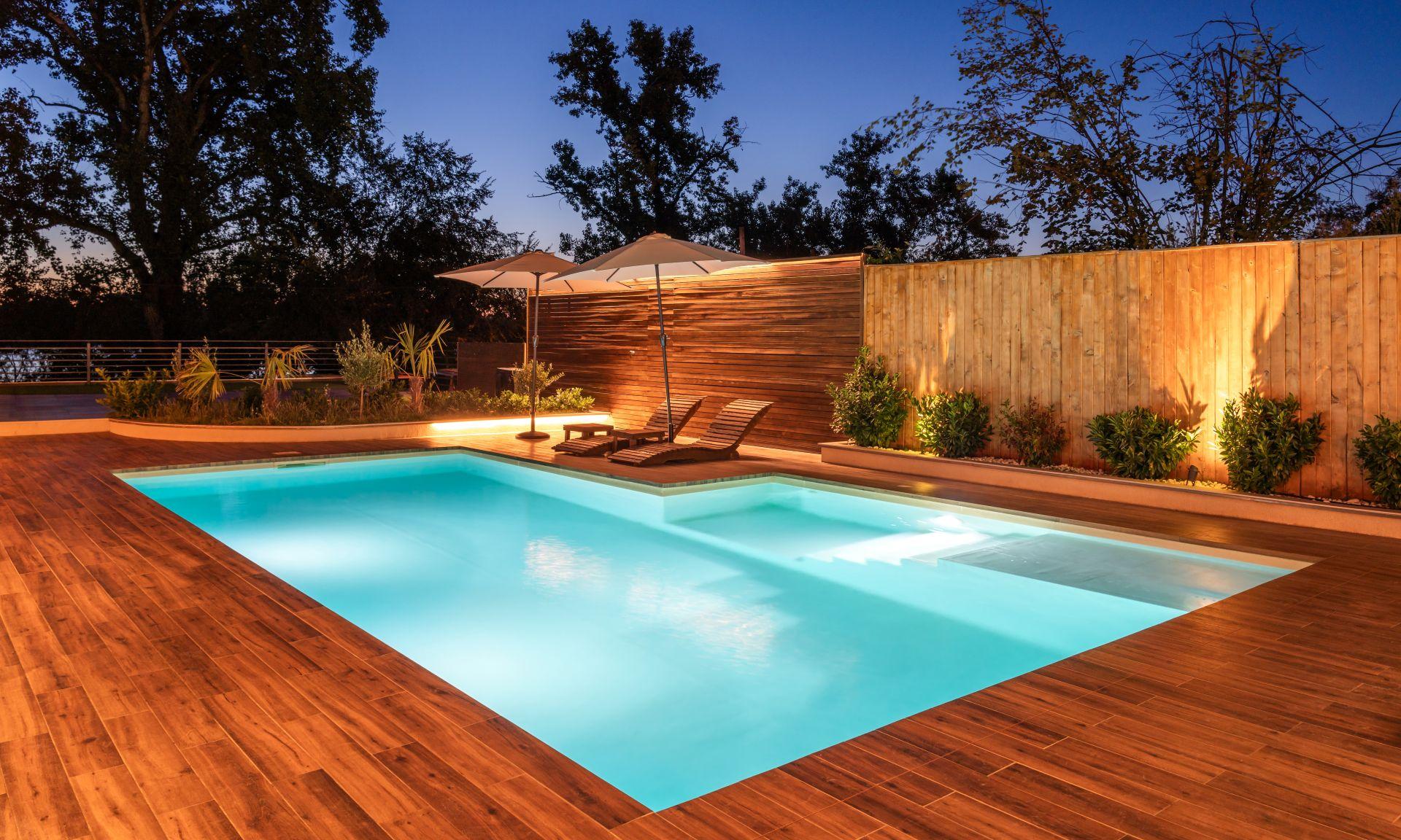 Beleuchteter Pool mit Holzdeck und zwei Sonnenliegen in einem ruhigen Garten bei Sonnenuntergang.